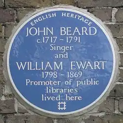 A Blue plaque on a brick wall with the words "John Beard C1717 – 1791 Singer and William Ewart 1798 – 1861 Promoter of Public Libraries