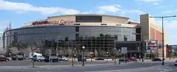 The wide, convex facade of a glassy sports arena and the parking lot, lawn, and lampposts in front of it. The words "Xcel Energy Center" sit in red atop the arena.